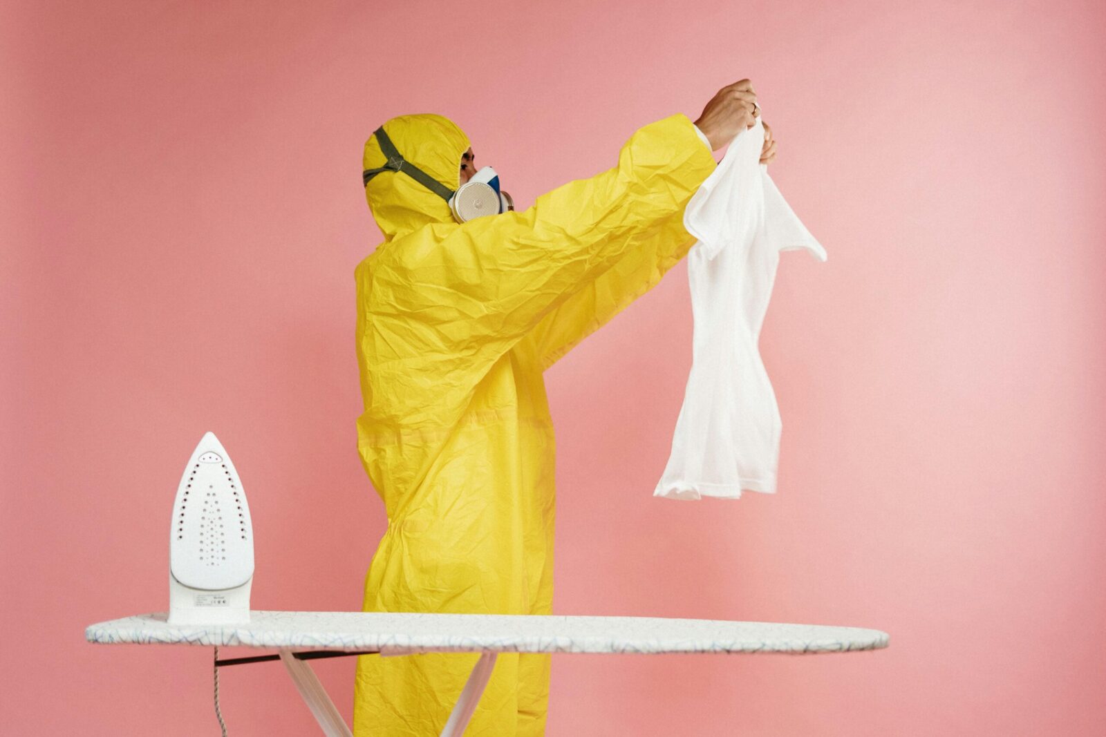 A man in yellow protective suit humorously ironing. Indoors on pink background.